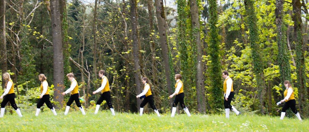 Sieben Personen in gelben und weißen Hemden, schwarzen Hosen und weißen Stiefeln gehen in einer Reihe auf einer Wiese mit Bäumen.