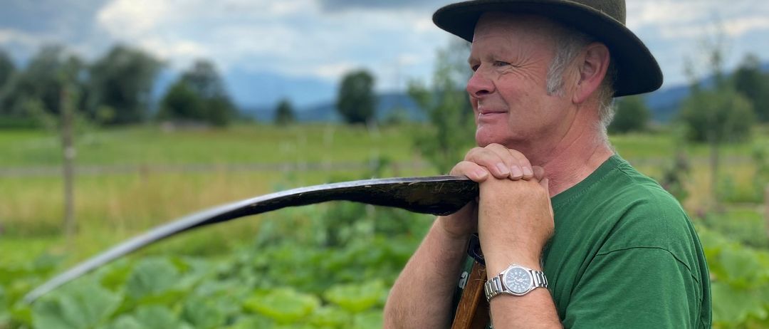 Ein älterer Mann in einem grünen Shirt und einer Mütze hält eine Sense in einem Feld mit bewölktem Himmel und Bergen im Hintergrund.