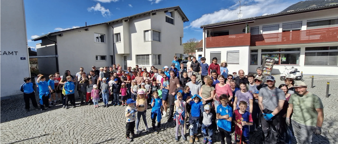 Eine große Gruppe von Menschen, darunter Erwachsene und Kinder, versammelt sich an einem sonnigen Tag vor einem weißen Gebäude. Einige halten Stöcke, und einige tragen Hüte und Brillen.