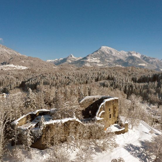 Eine Draufsicht auf eine schneebedeckte Ruine, umgeben von verschneiten Bäumen und Bergen unter einem klaren blauen Himmel.