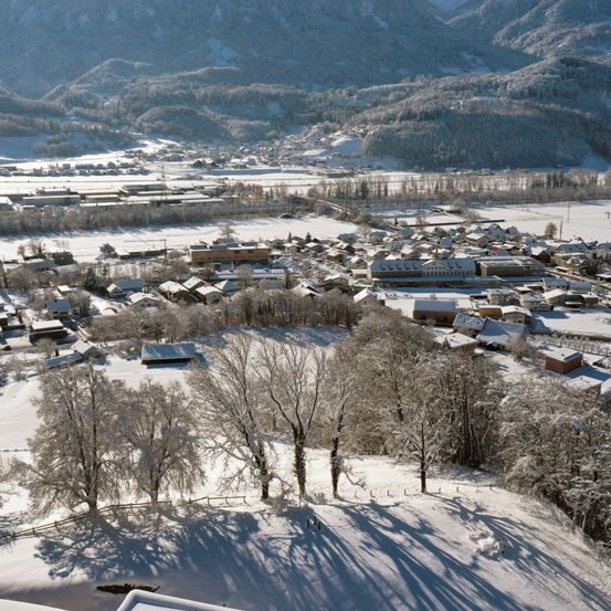 Ein verschneites Dorf umgeben von Bergen, mit Häusern und Bäumen, die in Schnee eingehüllt sind, und einem klaren blauen Himmel.