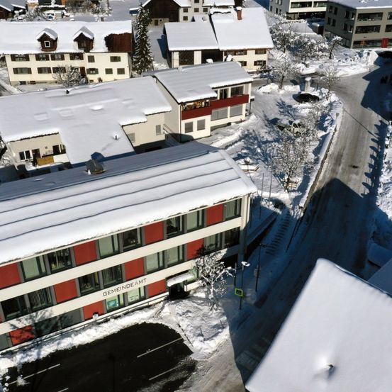 Luftaufnahme eines verschneiten Ortes mit einem Gebäude, das mit 'Gemeindeamt' beschriftet ist. Mehrere Gebäude, Straßen und Bäume sind zu sehen.