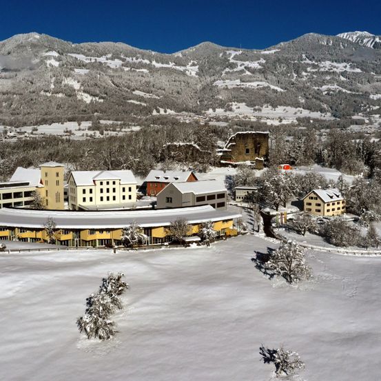 Eine Winterlandschaft mit einem Gebäudekomplex, umgeben von Schnee, Bergen im Hintergrund und schneebedeckten Bäumen.