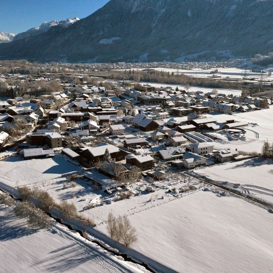 Luftaufnahme eines schneebedeckten Dorfes umgeben von verschneiten Bergen, mit Häusern und Gebäuden, die im Schnee vergraben sind, und einem Fluss, der durch die Landschaft führt.