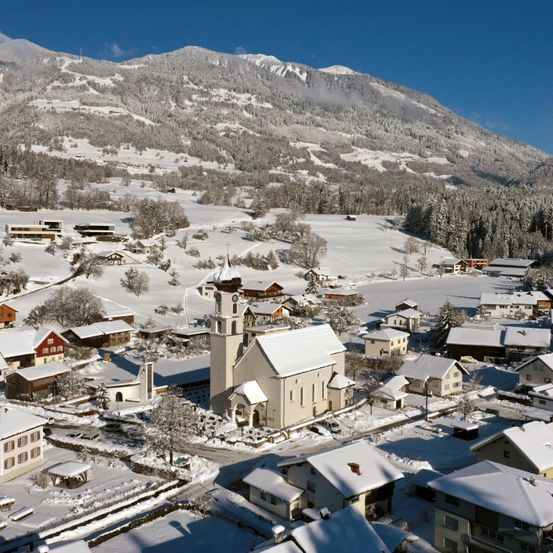 Luftaufnahme eines schneebedeckten Dorfes mit Häusern und einer Kirche, eingebettet in einem Tal, umgeben von verschneiten Bergen und Bäumen unter einem klaren blauen Himmel.