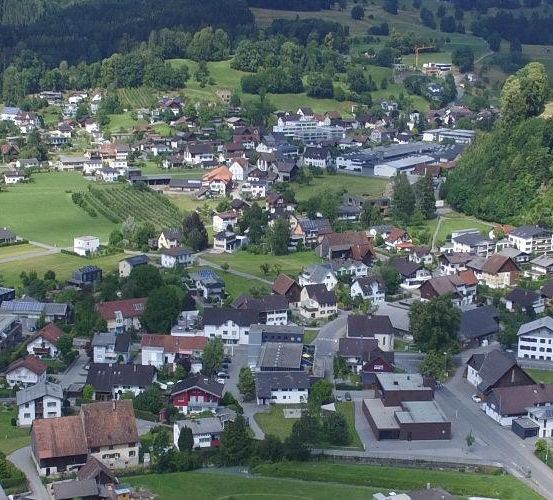 Luftaufnahme eines Dorfes mit vielen Häusern und Gebäuden, umgeben von grünen Feldern und Bäumen. Das Dorf liegt zwischen Bergen und hat eine Straße, die durch es führt.
