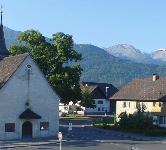 Eine kleine Kapelle mit einem Kirchturm steht an einer Straßenecke mit einem Stoppschild. Ein Gebäude mit Fenstern und einem Schornstein befindet sich auf der rechten Seite. Ein Auto ist vor einem anderen Gebäude geparkt. Bäume und Berge sind im Hintergrund.