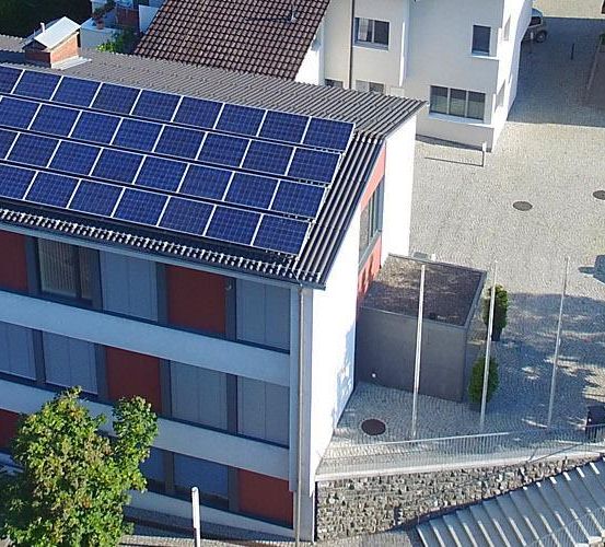 Ein Gebäude mit Solarmodulen auf dem Dach ist von oben zu sehen. Es ist von anderen Gebäuden, Bäumen und einem Parkplatz umgeben.