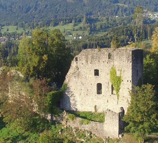 Ein Luftbild einer alten Steinburg, umgeben von Bäumen, mit einem Dorf im Hintergrund.