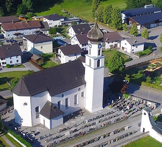 Eine Luftaufnahme eines kleinen Dorfes mit einer weißen Kirche und einem Friedhof mit vielen Grabsteinen.