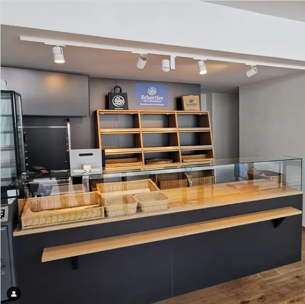 Ein Bäckereiladen-Interieur mit einer hölzernen Theke, die eine Glasvitrine, mehrere Weidenkörbe und ein großes blaues Schild mit der Aufschrift Schertler über einem Regal zeigt.