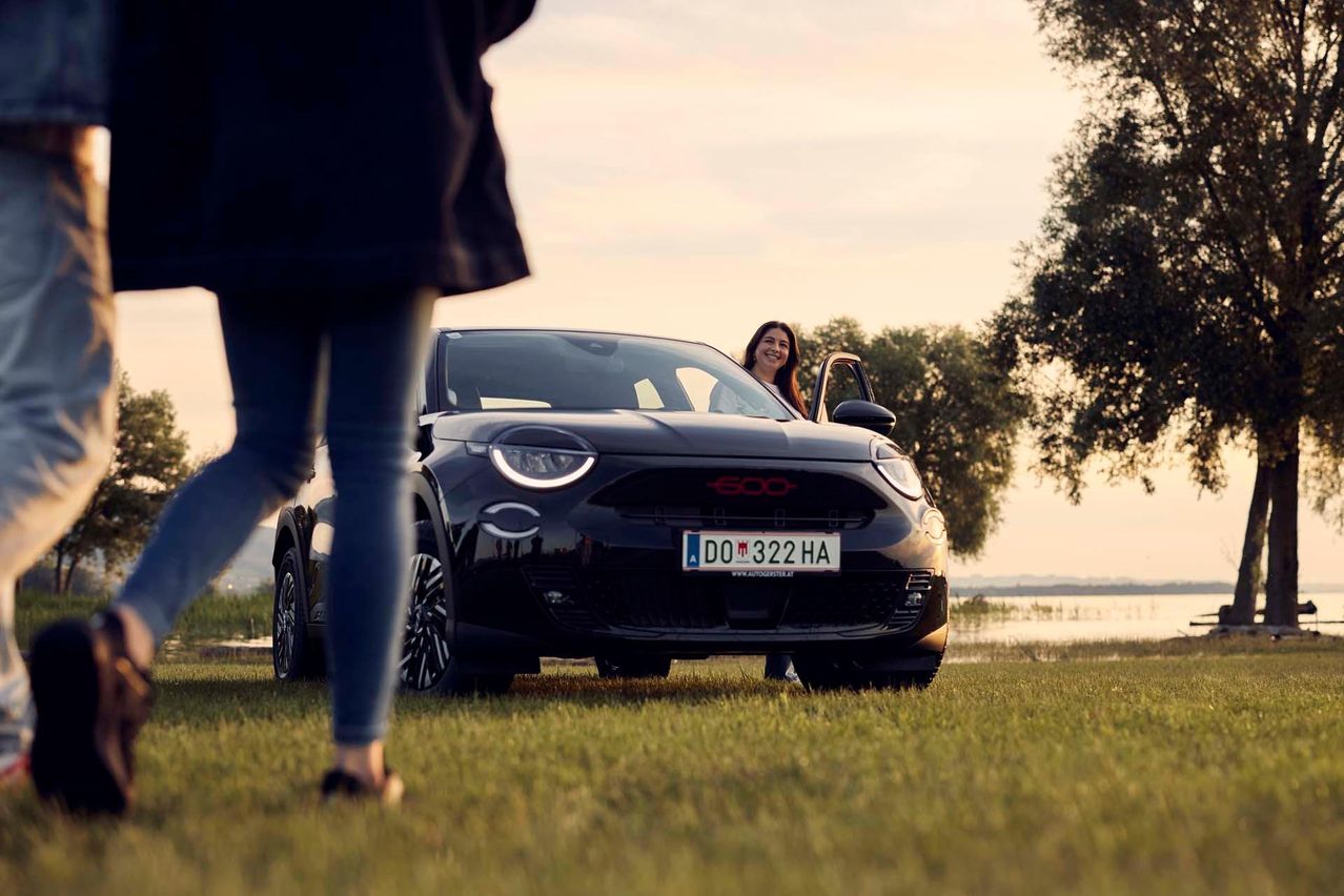 Eine Frau steht neben einem schwarzen umweltfreundlichen Auto, dessen Fahrertür geöffnet ist, lächelt und schaut in Richtung Kamera. Bäume und ein Gewässer sind im Hintergrund zu sehen.