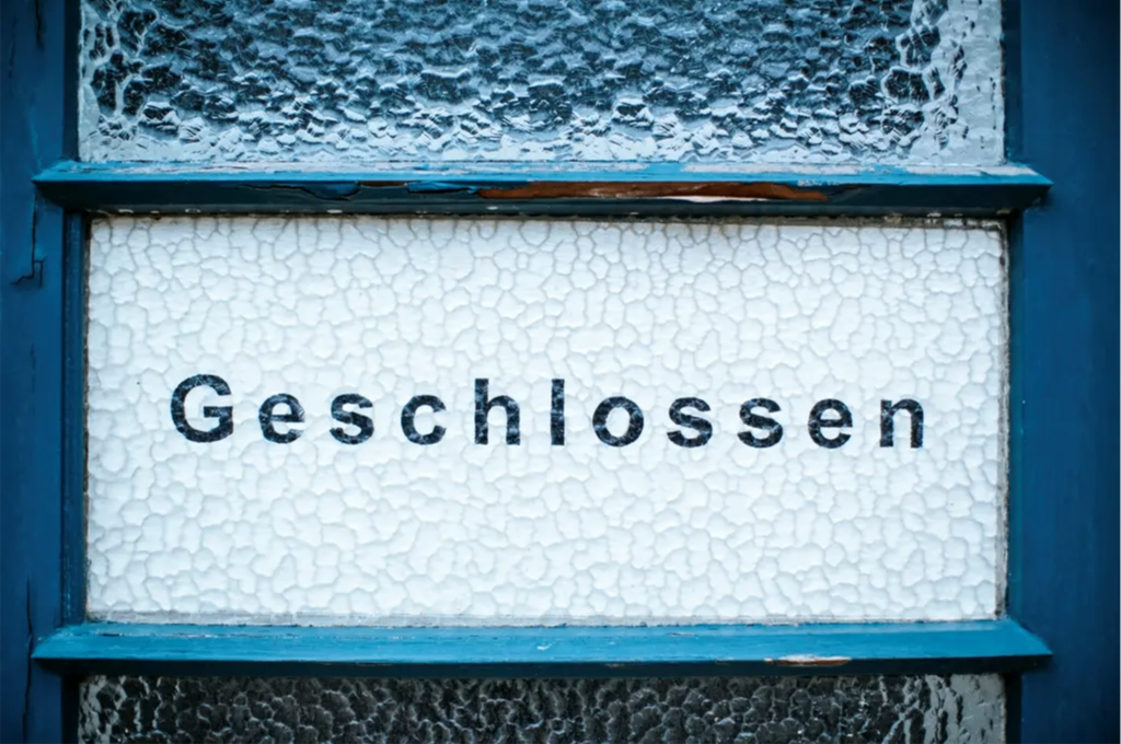 Ein weißes Schild mit blauem Rand zeigt das Wort 'Geschlossen' in fetten schwarzen Buchstaben auf einem strukturierten weißen Hintergrund.