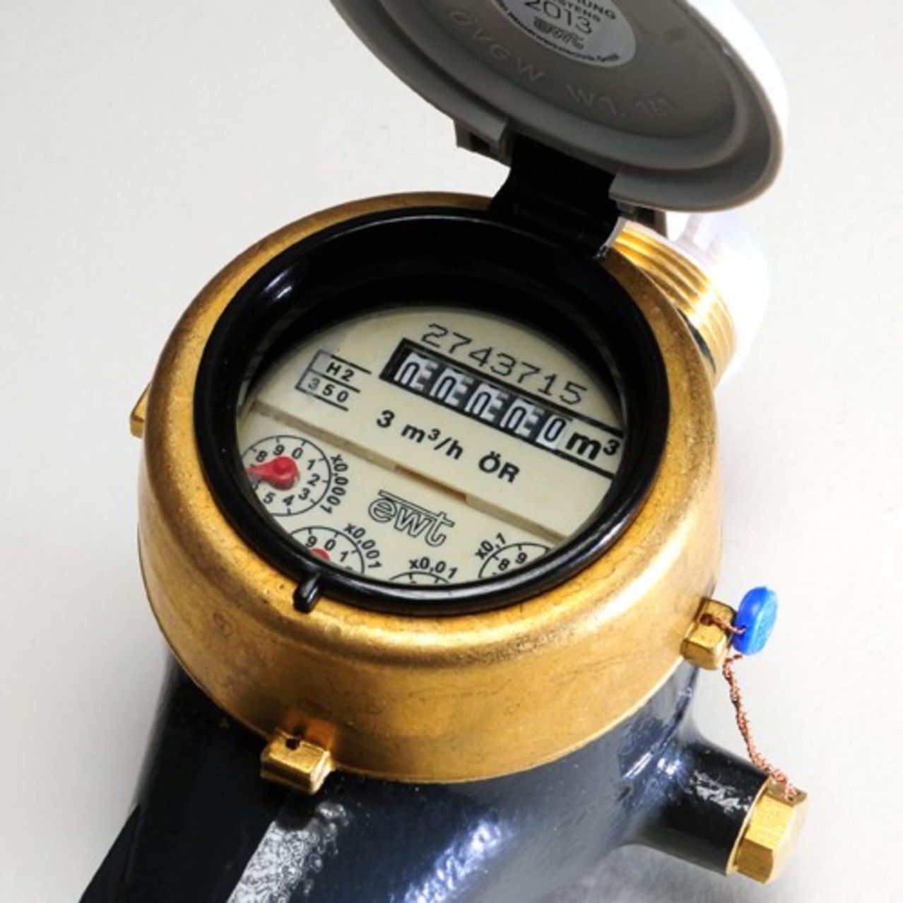 Ein geöffneter Wasserzähler mit einem goldfarbenen Körper und schwarzen Zifferblättern zeigt die Zahlen 2743715, 3 m3/h und 0 m3. Der Zähler hat einen blauen Knopf an der Seite.