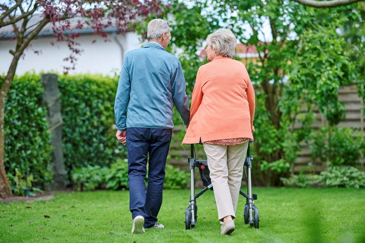 Ein älteres Paar geht Hand in Hand auf einem Rasen, die Frau benutzt einen Rollator. Dahinter befinden sich Bäume und Büsche.