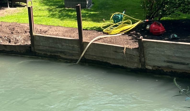 Ein Holzzaun begrenzt einen Teich, in den Wasser fließt, begleitet von einem gelben Schlauch und Gartengeräten.