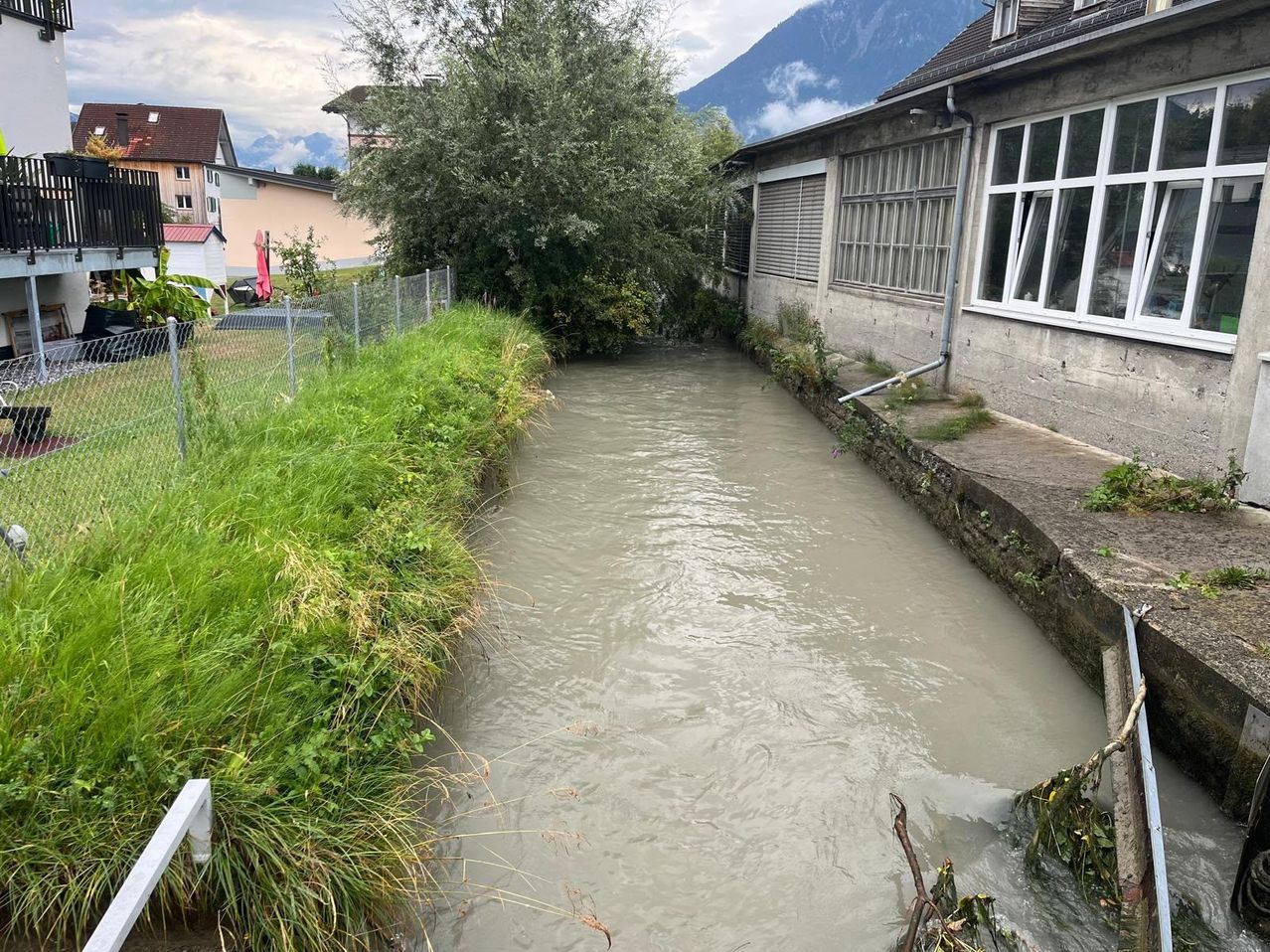 A muddy river with a concrete walkway on the right side, surrounded by grass and a fence. In the background, there are buildings and mountains.