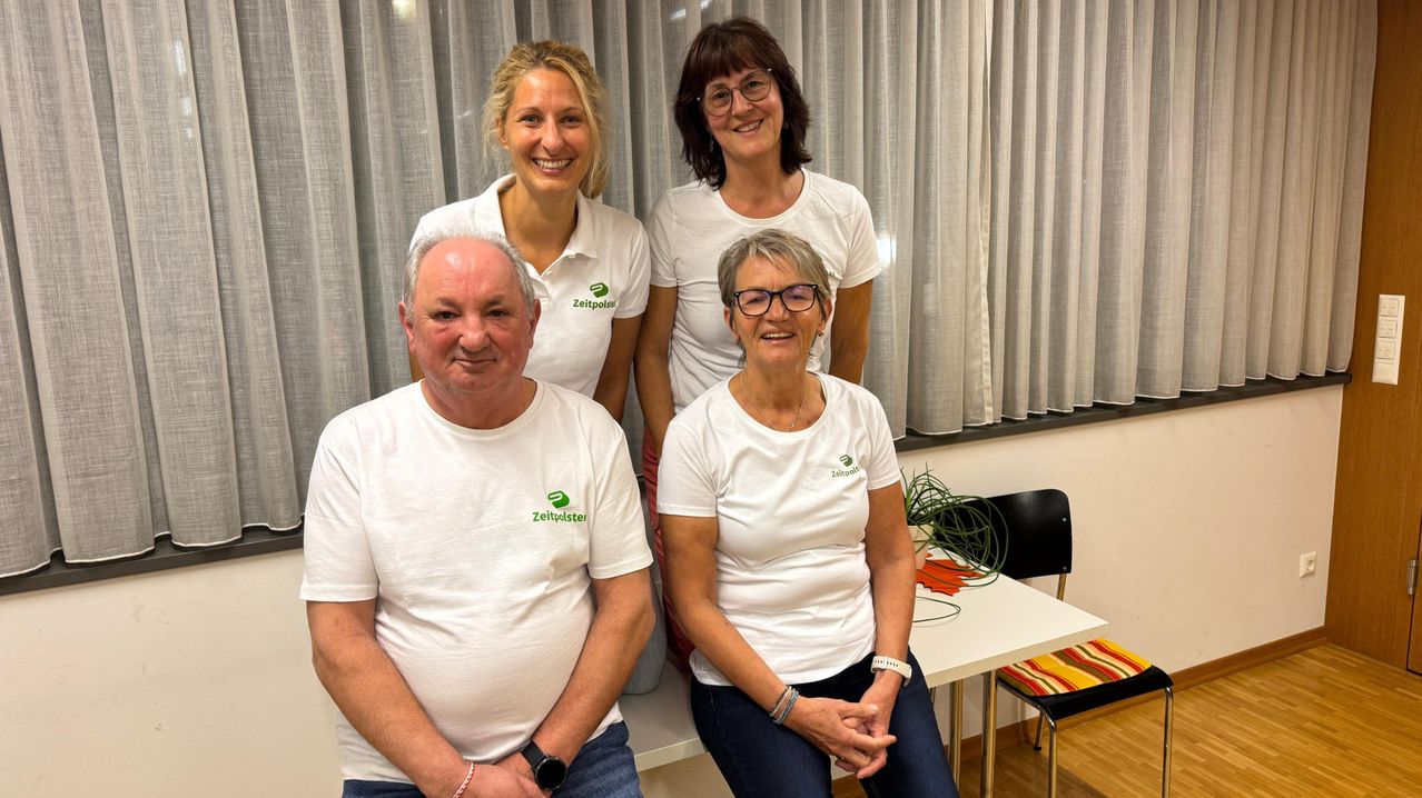 Vier Personen in weißen T-Shirts mit grünem Logo stehen lächelnd zusammen. Dahinter befindet sich ein weißer Vorhang. Es gibt einen Tisch mit einer Pflanze und einem schwarzen Stuhl.