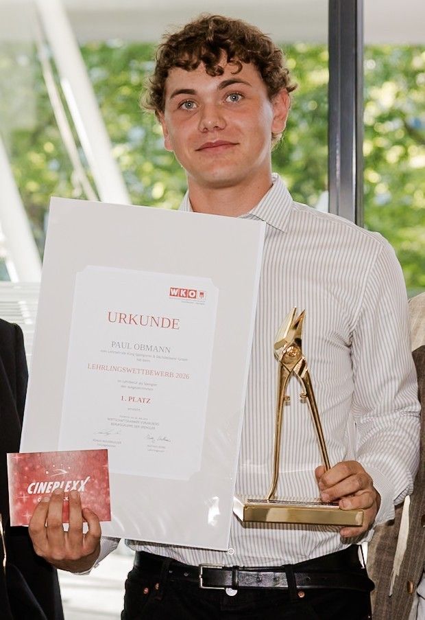 Ein Mann hält ein Zertifikat und eine Trophäe und lächelt. Er hat lockige Haare und trägt ein gestreiftes Hemd. Das Zertifikat besagt 'URKUNDE' und 'Lehrlingswettbewerb 2016'. Die Trophäe hat goldene Griffe.