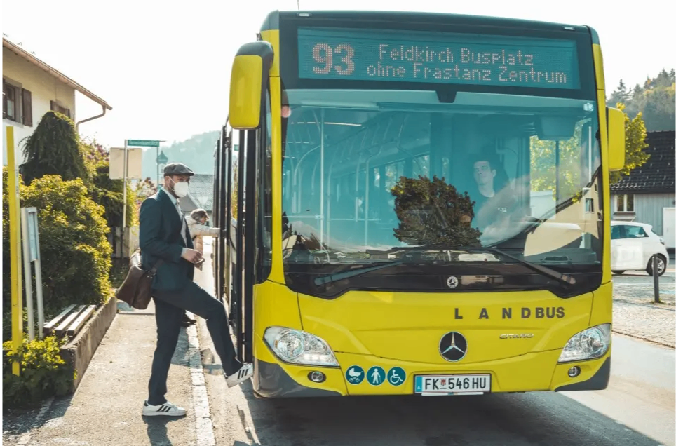 Ein Mann in Anzug und Maske steigt in einen gelben Land Bus ein. Der Bus zeigt ein digitales Schild mit der Aufschrift Feldkirch Busplatz ohne Fastanz Zentrum.