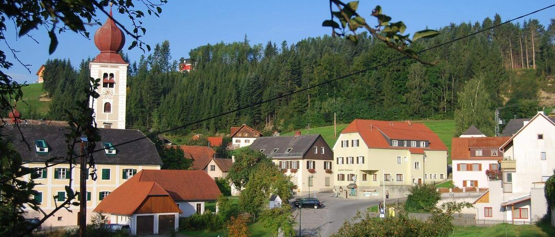 Luftaufnahme eines Dorfes in den Bayerischen Alpen mit einem Glockenturm, Häusern mit roten Dächern und einem klaren blauen Himmel.