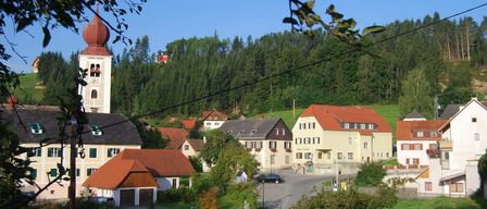 Luftaufnahme eines Dorfes in den Bayerischen Alpen mit einem Glockenturm, Häusern mit roten Dächern und einem klaren blauen Himmel.