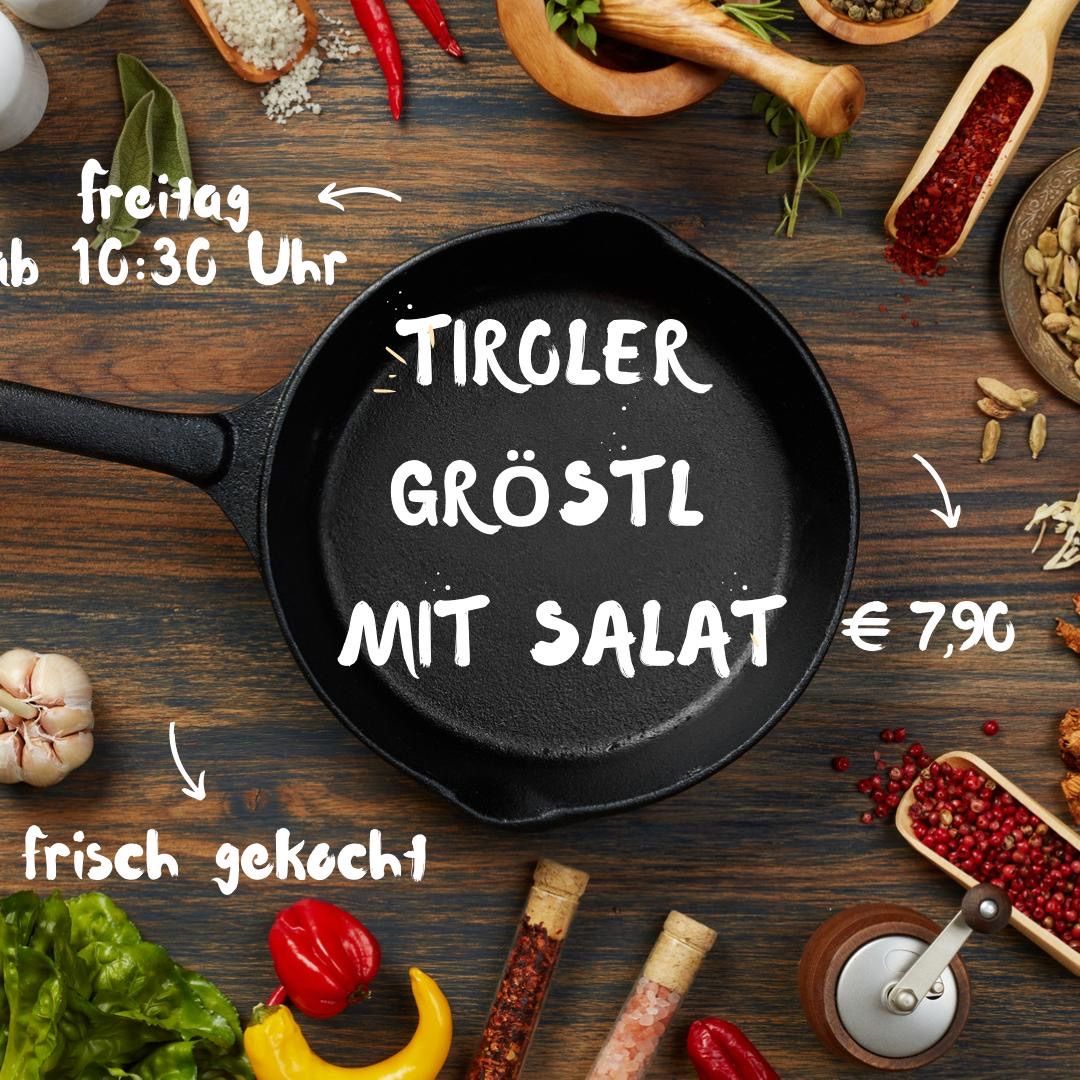 Eine schwarze Bratpfanne mit dem Text Tiroler Groestl mit Salat darauf. Die Pfanne steht auf einem Holztisch mit verschiedenen Zutaten und Küchenutensilien darum herum.