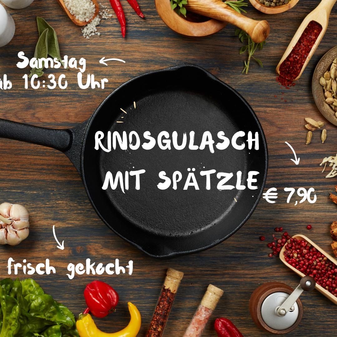 Auf einem Holztisch steht eine schwarze Bratpfanne mit der Aufschrift 'Rindsgulasch mit Spatzle'. Verschiedene Zutaten wie Knoblauch, rote Chilis und Gewürze sind herumgestreut. Der Preis ist mit 7,90 € angegeben. Das Bild enthält einen Zeitstempel für Samstag um 10:30 Uhr.
