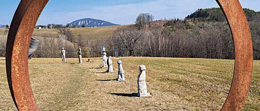 Ein kreisförmiges, verrostetes Metallgerüst umrahmt ein offenes Feld mit mehreren stehenden Figuren und einem entfernten Berg.
