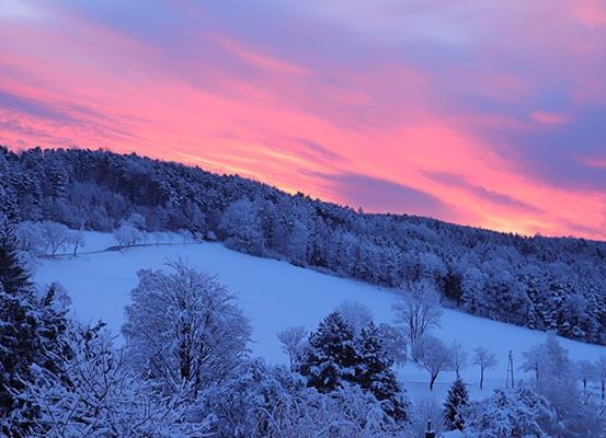 Eine verschneite Landschaft mit einem Gebirgszug, Bäumen und einem schneebedeckten Feld. Der Himmel ist rosa mit Wolken.