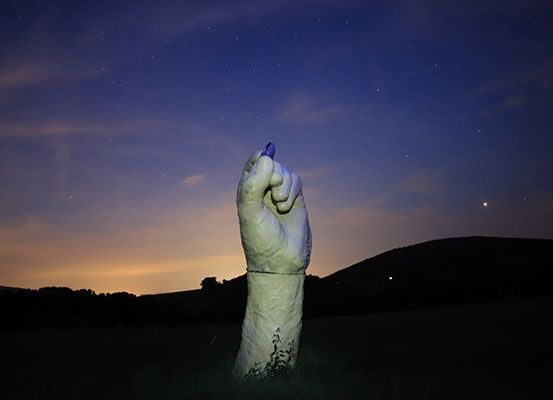 Eine große Handskulptur steht auf einem Feld unter einem sternenklaren Nachthimmel. Die Hand hat einen blauen Marker, der nach oben zeigt. In der Ferne sind Hügel und ein sonnenuntergangsfarbener Himmel zu sehen.