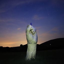 Eine große Handskulptur steht auf einem Feld unter einem sternenklaren Nachthimmel. Die Hand hat einen blauen Marker, der nach oben zeigt. In der Ferne sind Hügel und ein sonnenuntergangsfarbener Himmel zu sehen.