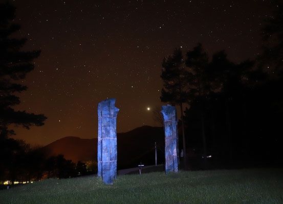 Zwei beleuchtete Steinsäulen stehen auf einem grasbewachsenen Hügel unter einem Sternenhimmel, mit Bergen im Hintergrund.