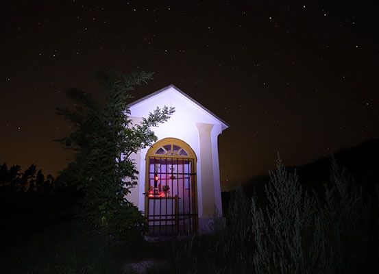 Eine kleine Kapelle auf dem Land, nachts von roten Lichtern beleuchtet, mit einem Tor und umgeben von Bäumen.