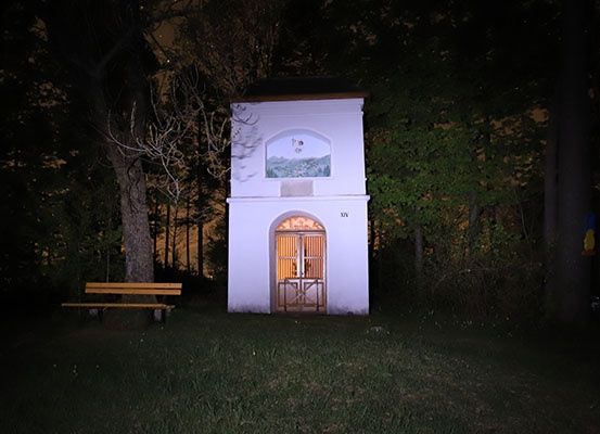 Eine kleine weiße Kapelle steht in einem dunklen, bewaldeten Bereich, mit einer Bank in der Nähe und Bäumen, die sie umgeben.