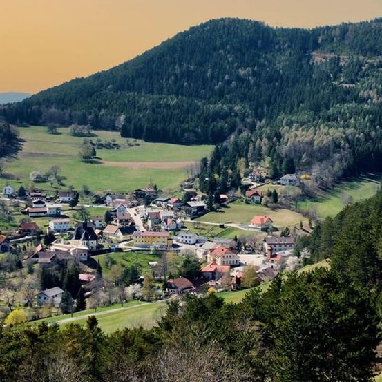 Luftaufnahme eines kleinen Dorfes, eingebettet in einem Tal, umgeben von grünen Feldern und Bäumen unter einem klaren Himmel.