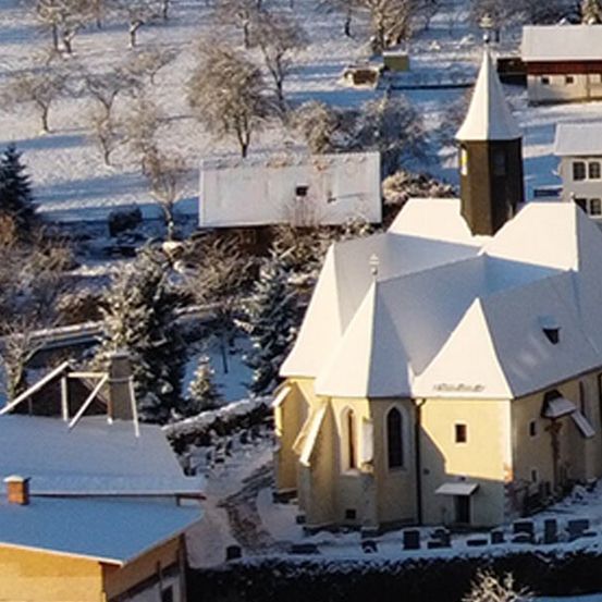 Luftaufnahme eines Dorfes mit einer schneebedeckten Kirche, umgeben von schneebedeckten Bäumen und Häusern in einer verschneiten Landschaft.