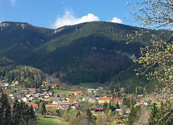 Ein Dorf in einem Tal unterhalb eines steilen Bergrückens. Bäume und Häuser sind sichtbar, mit einem klaren blauen Himmel darüber.