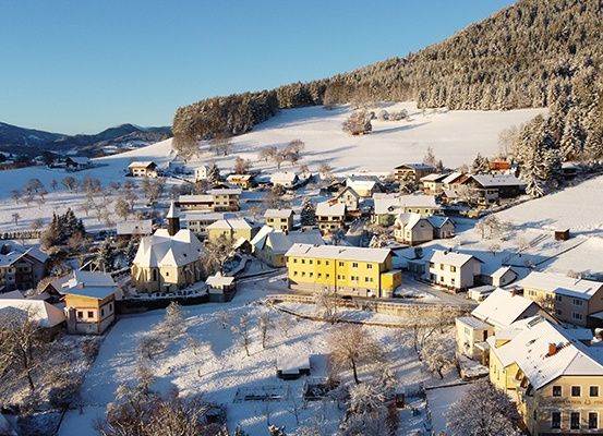 Ein von Schnee bedecktes Dorf mit Bergen im Hintergrund, Häusern und Bäumen sowie einer Kirche in der Mitte.