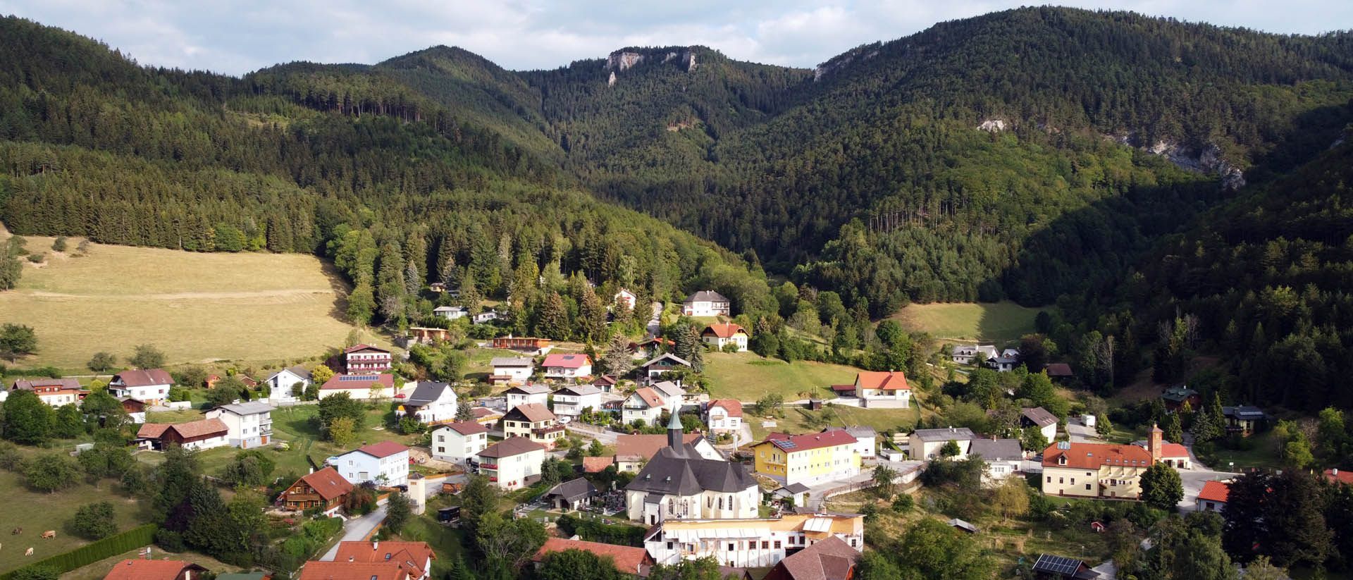 Luftaufnahme eines kleinen Dorfes in den Bergen mit üppigem Grün, Häusern und einer Kirche.