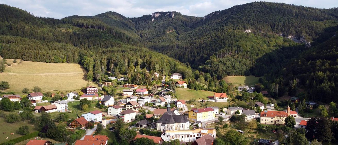 Luftaufnahme eines kleinen Dorfes in den Bergen mit üppigem Grün, Häusern und einer Kirche.