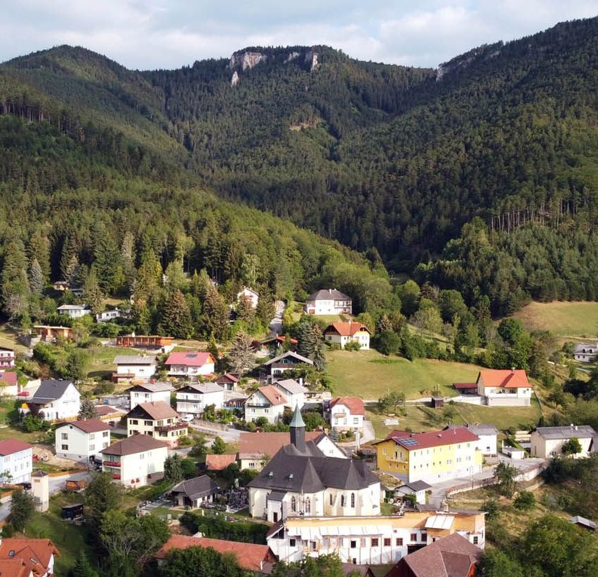 Luftaufnahme eines kleinen Dorfes in den Bergen mit üppigem Grün, Häusern und einer Kirche.
