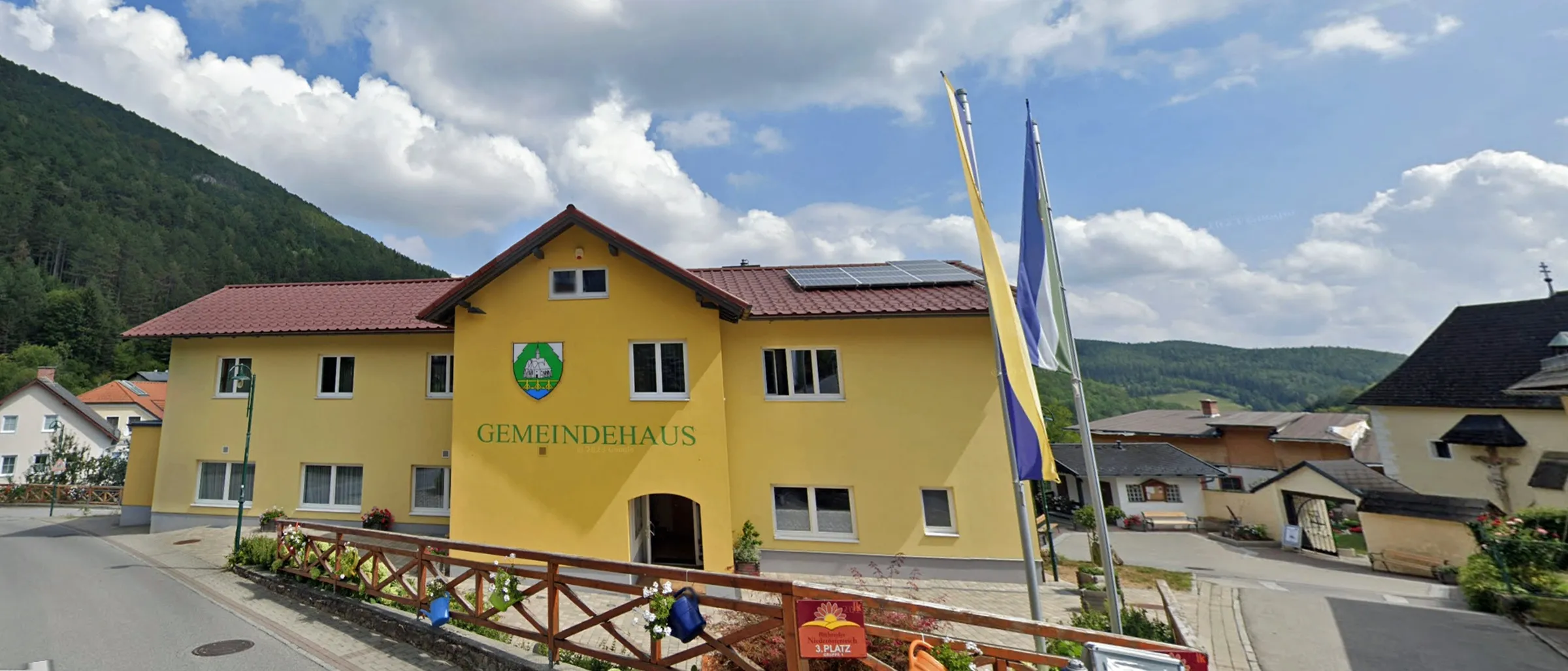 Ein gelbes Gebäude mit rotem Dach und Solarpanelen ist mit 'Gemeindehaus' beschriftet. Es ist von anderen Häusern und Bergen im Hintergrund umgeben.