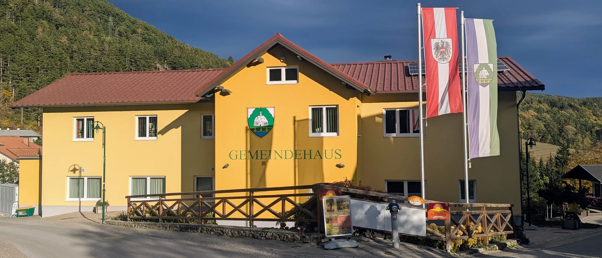 Ein gelbes Gebäude mit einem roten Dach und der Aufschrift 'Gemeindehaus'. Vor dem Gebäude stehen zwei Fahnen auf Stangen, und im Hintergrund befindet sich ein Berg.