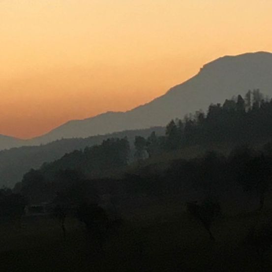 Eine Bergkette unter einem untergehenden Sonnenuntergang, mit silhouettierten Bäumen und einer entfernten Struktur. Der Himmel ist in Orange- und Rosatöne getaucht.