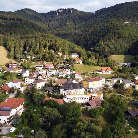 Luftaufnahme eines kleinen Dorfes, umgeben von grünen Hügeln und Wäldern, mit einer Kirche im Zentrum und Häusern verschiedener Größen und Farben.