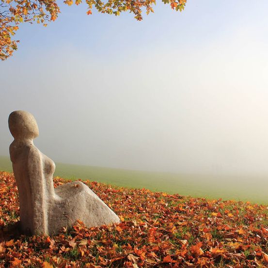 Eine Steinskulptur einer sitzenden Frau steht auf einem grasbewachsenen Hügel mit gefallenen Blättern. Der Himmel ist klar mit einem Hauch von Nebel.