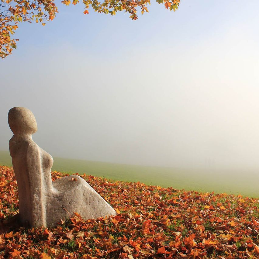Eine Steinskulptur einer sitzenden Frau steht auf einem grasbewachsenen Hügel mit gefallenen Blättern. Der Himmel ist klar mit einem Hauch von Nebel.