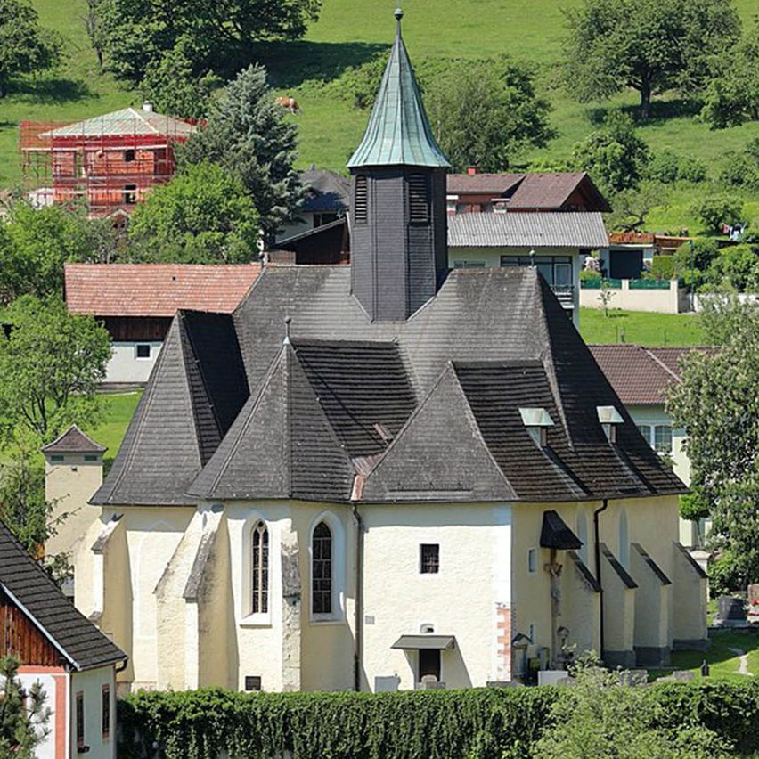 Eine Kirche mit einem Turm und einem Schieferdach steht in einem Dorf, umgeben von Bäumen und Hügeln. Mehrere Häuser sind im Hintergrund sichtbar.