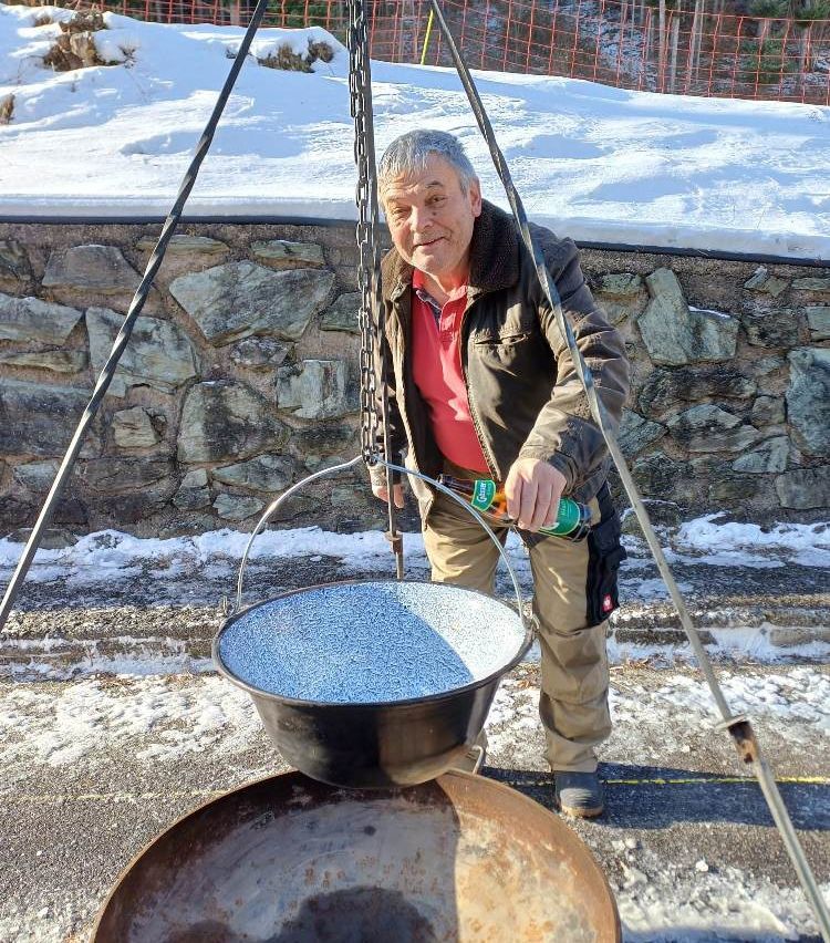Ein Mann steht in der Nähe einer schneebedeckten Mauer, hält eine Flasche und einen Topf mit einer Kette über einem Feuerstellen.